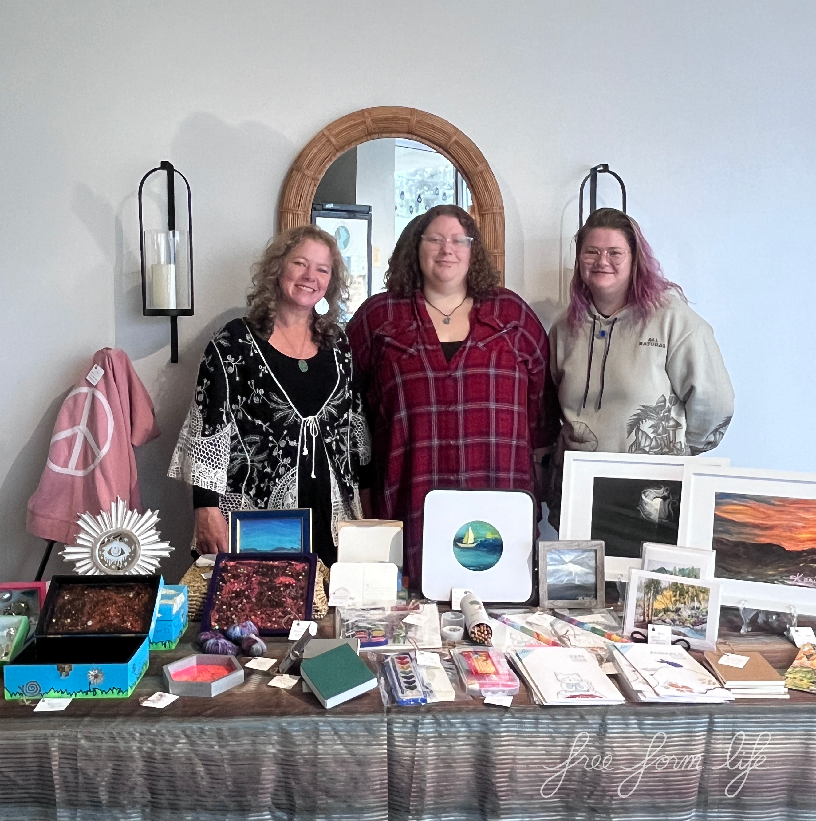 Three Free Form Life team members stand behind their pop-up shop event at the local yoga studio. The table is filled with art, prints, treasure boxes, resin trays, coloring books and more.