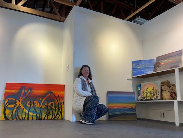 New studio with empty walls and artwork leaning against the wall.  Artist, Kariba, smiling in the center.